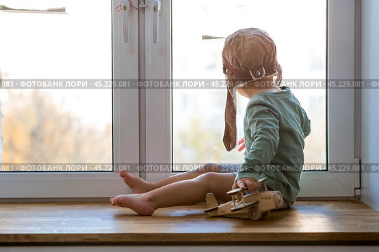 A cute 2-year-old boy in a cap is playing on the windowsill with a wooden airplane. Children's wooden toys for child development. Стоковое фото, фотограф Photojuli / Фотобанк Лори