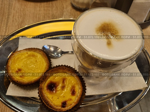 A delicious serving of latte coffee with thick milk foam and caramel pattern on the surface in a double glass mug. Next to it, on a metal tray, are two traditional Portuguese pastel de nata tartlets with a golden caramelized crust and delicate cream inside. Dessert and coffee are served on a white paper napkin with a small teaspoon. The warm wooden table and soft lighting create an atmosphere of coziness and relaxation, ideal for a coffee shop or home tea party. Стоковое фото, фотограф Анна Кривицкая / Фотобанк Лори
