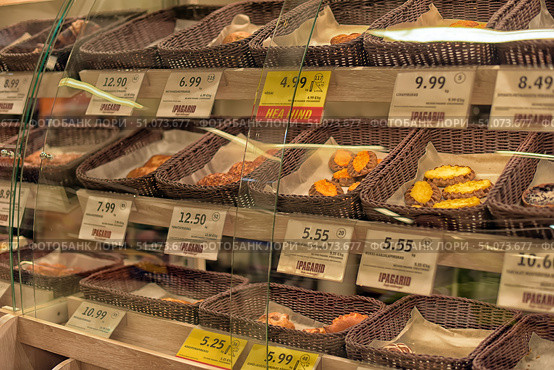 A display case with baked goods in a supermarket, divided by glass partitions. On the shelves are wicker baskets with various types of baked goods, pies and cookies. Each basket has a price tag indicating the cost per piece or kilogram. Among the assortment, you can see traditional products such as Karelian pies, cookies with curd filling and puff pastries. The price tags are in Estonian, and some products are highlighted with yellow price tags with discounts. The lighting is warm, emphasizing the golden color of the baked goods and creating a cozy atmosphere. (2015 год). Редакционное фото, фотограф Анна Кривицкая / Фотобанк Лори