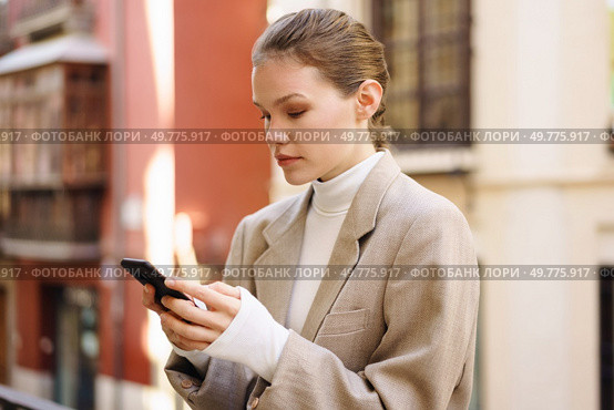 A fashionable woman in a beige blazer and cozy turtleneck engages with her smartphone in a vibrant urban setting, embodying modernity, elegance, grace, and poise with every interaction. A fashionable young woman skillfully using her smartphone while outdoors in daylight. Стоковое фото, агентство Ingram Publishing / Фотобанк Лори