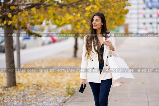 A fashionable woman strolls along a busy sidewalk, holding colorful shopping bags, surrounded by autumn foliage, showcasing her elegance and leisurely lifestyle with a joyful demeanor. A Stylish Woman Delightfully Enjoys Shopping on an Autumn Day Filled with Colorful Leaves. Стоковое фото, агентство Ingram Publishing / Фотобанк Лори