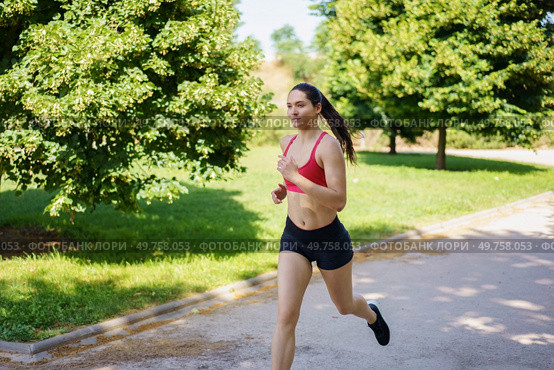 A fit woman in sportswear is jogging in a lush green park, enjoying the sunny day, maintaining a healthy lifestyle, and staying active to keep her vitality and wellness at peak levels. A young woman is jogging in the park on a sunny day, enjoying her workout in the fresh air surrounded by nature. Стоковое фото, агентство Ingram Publishing / Фотобанк Лори