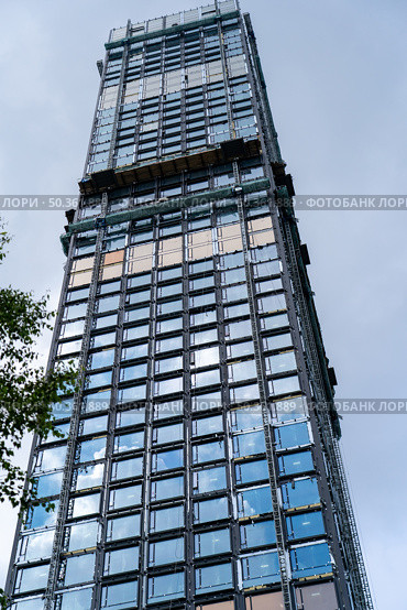 A high-rise building is being constructed, showcasing a facade of glass panels and metal supports against the blue sky. Стоковое фото, фотограф Данила Большаков / Фотобанк Лори