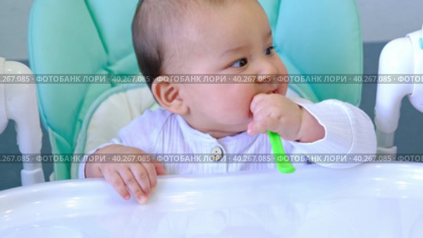 A hungry baby is gnawing on a plastic spoon at the table on a high chair. Teething, whims, itchy gums, introduction of complementary foods. Стоковое видео, видеограф Ольга Симонова / Фотобанк Лори