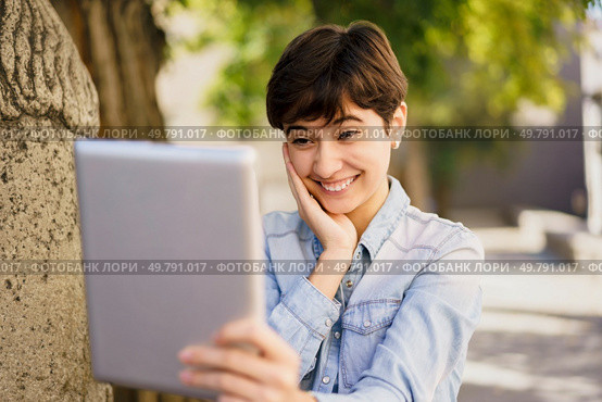 A joyful, bright, and carefree young woman is having a lively video call outdoors, smiling widely while holding a tablet in her hands, surrounded by the beauty of nature all around her. A Young Woman Happily Enjoying a Video Call Outdoors Using Her Tablet in the Sunshine. Стоковое фото, агентство Ingram Publishing / Фотобанк Лори