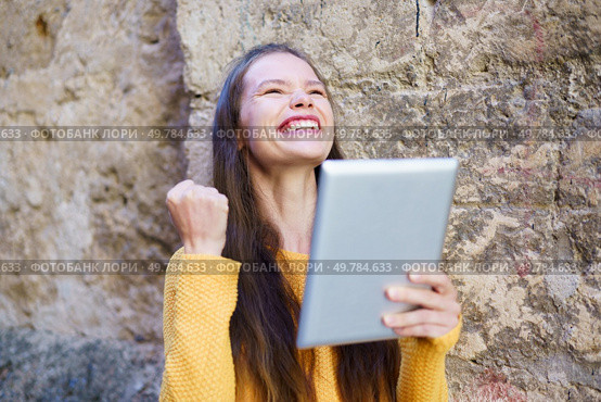 A joyful young woman celebrates her remarkable success while proudly holding up a tablet, expressing her excitement and happiness against the eyecatching backdrop of a rustic wall. A Joyful Young Woman is Celebrating with a Tablet in Her Hand, Radiating Happiness and Enthusiasm. Стоковое фото, агентство Ingram Publishing / Фотобанк Лори