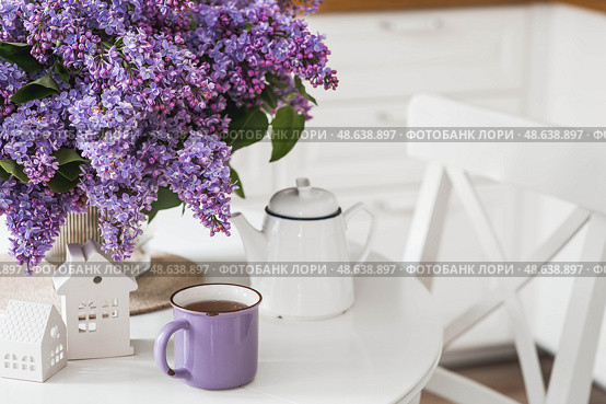 A large purple bouquet of lilacs, white Scandinavian houses, a ceramic teapot and a cup on the table. In the background is the interior of a modern white kitchen. The concept of home comfort. Стоковое фото, фотограф Photojuli / Фотобанк Лори