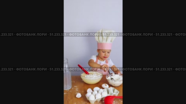 A little girl in a chef's hat and apron pours flour from a bowl into a dough with a spoon. On the table are a whisk, eggs, flour. Стоковое видео, видеограф Anna Scherbina / Фотобанк Лори