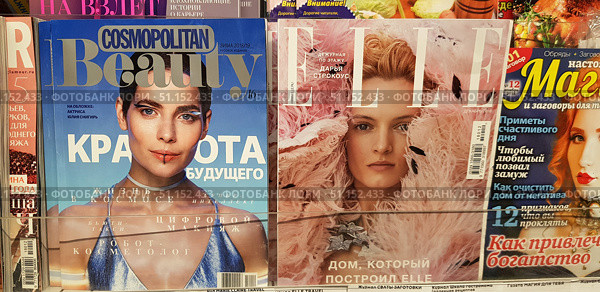 A magazine display case in a store. Three covers are clearly visible in the foreground: Cosmopolitan Beauty (winter 2018/19) with a portrait of actress Yulia Snigir, ELLE magazine (December 2018) with model Daria Strokous, and Magic and Conspiracies magazine with a bright cover promising tips on attracting wealth and amulets against negativity. The covers are made in different styles: a glossy fashion magazine with futuristic accents, a fashion publication with an airy image of a model, and an esoteric magazine with catchy headlines. The photo conveys the atmosphere of a kiosk or a printed media store, a variety of styles and content of modern periodicals. Редакционное фото, фотограф Анна Кривицкая / Фотобанк Лори