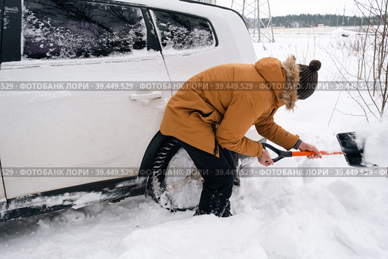 A man digs out a stalled car in the snow with a car shovel. Transport in winter got stuck in a snowdrift after a snowfall, sat on the bottom. First aid, tow truck, winter tires spikes and all-season. Стоковое фото, фотограф Ольга Симонова / Фотобанк Лори