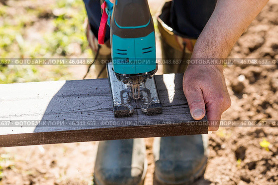 A man holds a battery-powered electric saw and builds new beds in a greenhouse made of wood-polymer composite. Macro photography. Side view. Preparing for the spring garden season. Стоковое фото, фотограф Photojuli / Фотобанк Лори