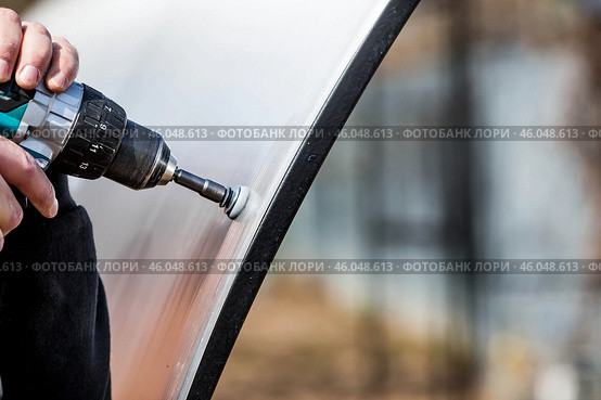 A man holds a battery-powered screwdriver and assembles a new greenhouse made of polyethylene, polycarbonate. Macro photography. Side view. Preparing for the spring garden season. Стоковое фото, фотограф Photojuli / Фотобанк Лори