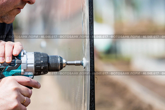 A man holds a battery-powered screwdriver and assembles a new greenhouse made of polyethylene, polycarbonate. Macro photography. Side view. Preparing for the spring garden season. Стоковое фото, фотограф Photojuli / Фотобанк Лори