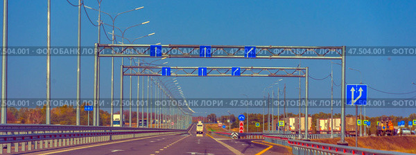 A multi-lane section of highway with perfectly smooth asphalt and metal guardrails. Above the roadway, structures with blue road signs are installed, indicating the directions of movement along the lanes. Light poles with identical curved consoles are built in perspective along the road, creating a rhythmic visual effect. On the right side of the frame is an exit from the highway leading to a parking lot and roadside infrastructure. Cars and trucks are visible in the distance. On the left is an autumn forest and a sign with the mileage. (2020 год). Редакционное фото, фотограф Анна Кривицкая / Фотобанк Лори