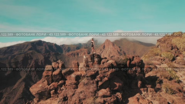 A person perches on a cliff edge with a panoramic view of mountainous terrain. Woman surveys the sprawling wilds, the peaks rising over cloudy sky. Стоковое видео, видеограф Ольга Сухарева / Фотобанк Лори