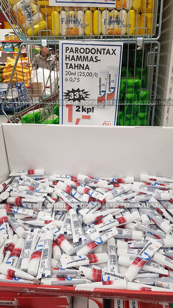 A photo from a supermarket shelf, where Parodontax toothpaste is presented in small 20 ml tubes. The tubes are laid out in a large box, above which there is an advertising poster with information about a -33% discount and the price for the package. Other products are visible in the background: laundry soap, food and customers. The image conveys the atmosphere of the sales area, focusing on the promotion and affordable price of the popular brand of toothpaste. (2019 год). Редакционное фото, фотограф Анна Кривицкая / Фотобанк Лори