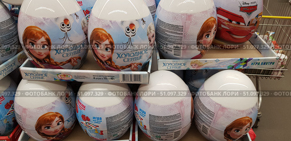 A photo of a store display case with large plastic surprise eggs featuring characters from the Disney cartoon Frozen. The packaging features Anna, Elsa, and the snowman Olaf on a blue and pink background with icy patterns. The eggs contain marmalade and a board game for the whole family, as indicated on the packaging. The inscription Frozen is visible in the center, and the Disney logo is on the side. In the background, you can see eggs featuring characters from the cartoon Cars. The bright and colorful children's products are presented in boxes and are intended as gifts for children. (2018 год). Редакционное фото, фотограф Анна Кривицкая / Фотобанк Лори