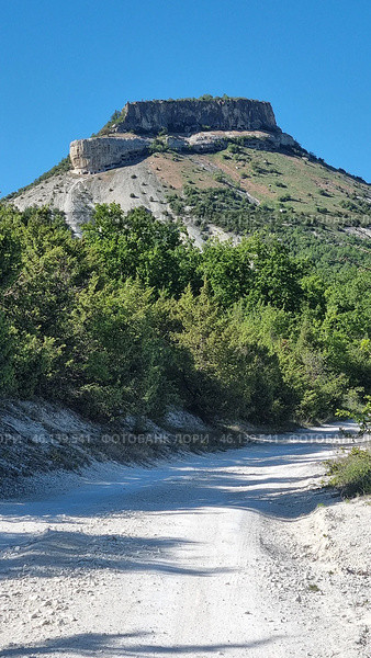 A picturesque Crimean landscape with a dirt road leading to a massive flat-topped rock. This mountain features steep cliffs and layered limestone rocks, which is typical of many Crimean mountain ranges. The lower part of the mountain is covered with dense green trees and bushes, creating a contrast with the dry, rocky slope.Mountain definition:The image probably depicts Mount Kyz-Kermen, one of the notable Crimean flat-topped mountains located near Bakhchisaray. This is an ancient cave settlement, used in the Middle Ages as a fortified settlement. The mountain has expressive rock ledges and grottoes, which can be seen in the photograph. Стоковое фото, фотограф Анна Кривицкая / Фотобанк Лори