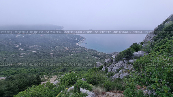 A picturesque view from the Laspi observation deck, located on the southern coast of Crimea. From the heights, a panorama of the coastline stretching into the distance in a light haze opens up. In the foreground, massive rocks, green bushes and mountain grasses are visible. Below, dense forests stretch out, turning into a coastal zone with houses, roads and piers. The sea is calm, bluish-gray, with ships visible on the horizon. The atmosphere is filled with misty softness and a sense of space, and the shooting point itself emphasizes the grandeur of Crimean nature and the harmony of the mountain and sea landscape. Стоковое фото, фотограф Анна Кривицкая / Фотобанк Лори