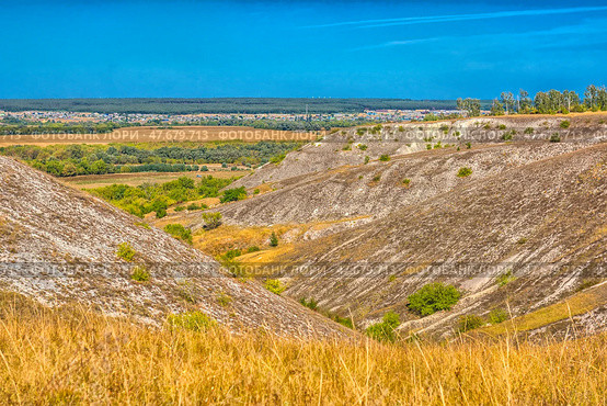 A picturesque view of chalk hills covered with dry steppe vegetation. In the foreground is tall dry grass, typical of the southern Russian steppe. Whitish stripes of chalk and sparse bushes are visible on the slopes of the hills. Below is a green valley with trees, behind it are yellow fields and a village. On the horizon, a forest and a light haze are visible. The bright blue sky without clouds emphasizes the contrast between the hills and the plain. This is probably one of the natural landscapes of the Voronezh region, for example, Divnogorye. Стоковое фото, фотограф Анна Кривицкая / Фотобанк Лори