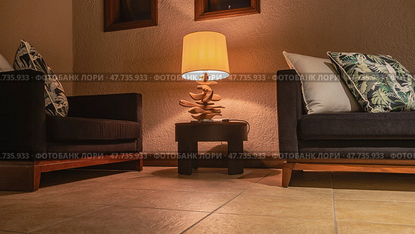 A place to relax in the room. Two sofas with soft patterned cushions against a beige wall in the corner. The desk lamp is lit. Translucent lampshade, designer wooden stand. Evening lighting. Стоковое фото, фотограф Вера Ивановна Кубарева / Фотобанк Лори