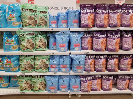 A rack in a pet store or supermarket with a large selection of cat litter. The shelves are lined with packages of different brands and colors. On the left are blue 8-liter Pretty Cat packages, in the center are light green 6-liter Cat Step Tofu Green Tea packages and blue CC Cat packages, on the right are purple Cat Step packages with a picture of a ginger cat. The packages are neatly arranged in rows, with price tags under each shelf: from 308 to 512 rubles, depending on the brand and volume. The photo demonstrates the variety of products and the order of their display in the store. (2019 год). Редакционное фото, фотограф Анна Кривицкая / Фотобанк Лори