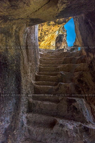 A rock-cut staircase leading up to daylight. The stone steps are uneven, rough, carved by time and probably by human hands. The light pours in from above, creating a beautiful play of shadows and emphasizing the texture of the ancient stone. Such a scene is typical of the cave monasteries of Crimea, especially Chelter-Marmara, one of the most atmospheric cave complexes, located near Bakhchisaray. It was founded in the Middle Ages and was used as a monastery. (2021 год). Стоковое фото, фотограф Анна Кривицкая / Фотобанк Лори