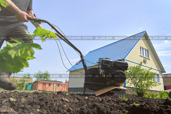 A rural neighbor is working in their garden, using a tiller to till the soil on a bright May day. Стоковое фото, фотограф Скрипачев Антон / Фотобанк Лори