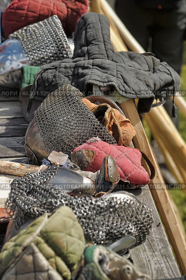 A set of medieval armor consisting of various protective elements. The focus is on the chainmail hood (aventail), covering the knight's helmet bascinet. Nearby are padded textile gloves with plate inserts, used to protect the hands in battle. Also present is a gambyson - a quilted jacket that serves as underarmor clothing, cushioning blows.In the frame, fragments of a shield, chain mail, parts of plate armor and a wooden stick, possibly used as a training weapon, are visible. This set of equipment is clearly intended for participation in historical reconstructions of medieval battles or knightly tournaments. Стоковое фото, фотограф Анна Кривицкая / Фотобанк Лори
