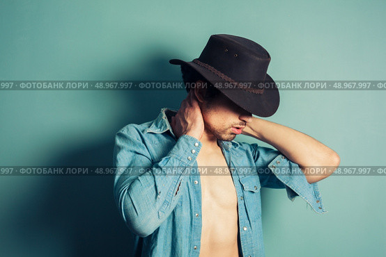 A sexy young cowboy is standing by a blue wall with his shirt unbuttoned. Стоковое фото, агентство Ingram Publishing / Фотобанк Лори