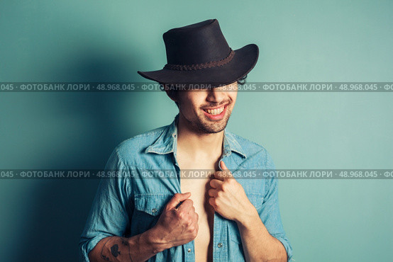 A sexy young cowboy is standing by a blue wall with his shirt unbuttoned. Стоковое фото, агентство Ingram Publishing / Фотобанк Лори