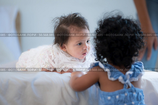 A small girl stood at the foot of the bed playing with the younger sibling carried by his mother and smiling with affection, In a bedroom surrounded by warmth and family. Стоковое фото, агентство Ingram Publishing / Фотобанк Лори