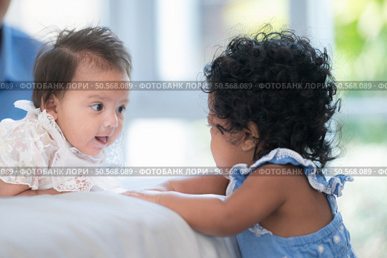 A small girl stood at the foot of the bed playing with the younger sibling carried by his mother and smiling with affection, In a bedroom surrounded by warmth and family. Стоковое фото, агентство Ingram Publishing / Фотобанк Лори