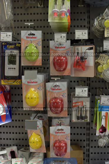 A stand in a store with kitchen accessories. In the center of the display hang mechanical kitchen timers from the Metaltex brand in the shape of fruits: apples (red and green) and pears (yellow). Each timer is designed for 60 minutes and is packed in a branded blister. Nearby you can see various kitchen products: bottle caps, a thermometer, wine accessories and metal tea strainers. All the products are neatly hung on hooks on the stand, with price tags above them. The photo clearly demonstrates the assortment of the hardware store and useful little things for the kitchen. (2015 год). Редакционное фото, фотограф Анна Кривицкая / Фотобанк Лори