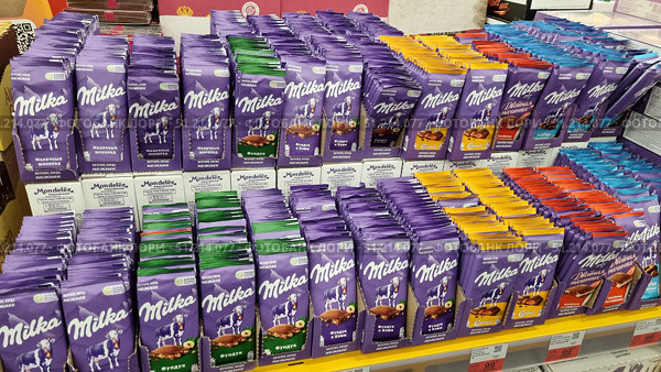A store counter with a large selection of Milka chocolate bars. The chocolate bars in characteristic purple packaging are laid out in neat rows. The packaging depicts the signature Milka cow against the backdrop of the Alpine mountains. Various flavors are visible: milk chocolate, chocolate with hazelnuts, with raisins and nuts, with caramel, with double filling, and also with air bubbles. Bright colored inserts on the packaging (green, yellow, red, blue) emphasize the variety of the assortment. The photo conveys the atmosphere of a grocery store, the abundance of sweets and the attractive display of the chocolate brand well. Редакционное фото, фотограф Анна Кривицкая / Фотобанк Лори