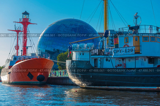 A striking and recognizable scene in Kaliningrad is the museum complex on the embankment. In the foreground are two ships: a red lighthouse ship and a black and white fishing vessel "CPT-129". In the center of the frame, against the backdrop of the ships, rises a spherical building - this is the Museum of the World Ocean with its famous dome-planetarium, lined with mirrored glass. Around - modern urban architecture, water and a clear blue sky. (2020 год). Редакционное фото, фотограф Анна Кривицкая / Фотобанк Лори