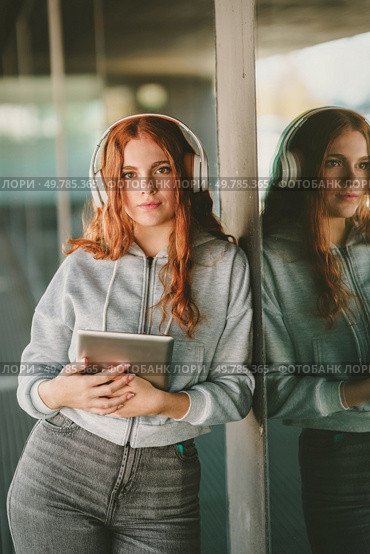 A stylish young woman with long red hair and modern headphones leans against a pillar holding a trendy tablet, embodying creativity and vibrancy in urban life through technology and individuality. A Young Woman Wearing Headphones and Engaging with a Tablet While in a Vibrant Urban Setting. Стоковое фото, агентство Ingram Publishing / Фотобанк Лори
