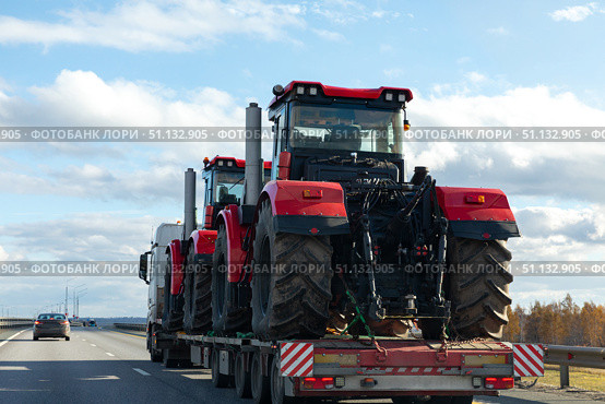 A tow truck transports two red farm tractors along a highway after work in field. Heavy-duty transport and agricultural machinery delivery concept. Logistics cargo transport. Стоковое фото, фотограф Ирина Михайличенко / Фотобанк Лори