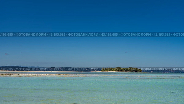 A tropical island covered with green vegetation and a sandbar are visible in the aquamarine ocean. Tiny silhouettes of people on a sandy beach. Mountains on the horizon. Clear blue sky. Copy space. Стоковое фото, фотограф Вера Ивановна Кубарева / Фотобанк Лори