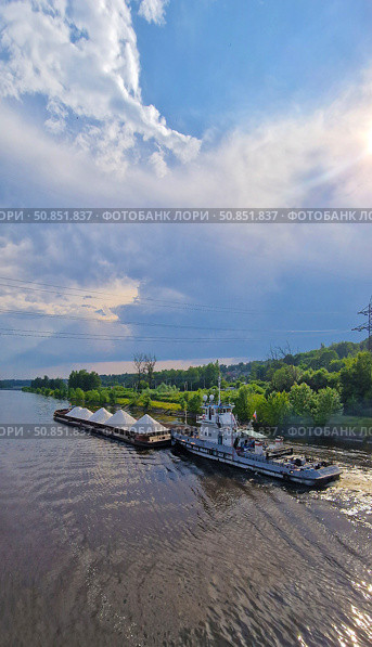 A tugboat pushes a barge with white rubble along a calm river. There are green trees and woodlands in the background, under a clear summer sky with thin clouds. Стоковое фото, фотограф Анна Чижова / Фотобанк Лори