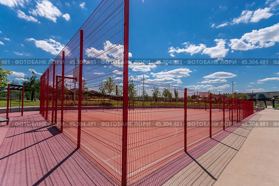 A vibrant red sports court surrounded by fencing in a sunny park setting on a clear day. Стоковое фото, фотограф Алексей Климкин / Фотобанк Лори