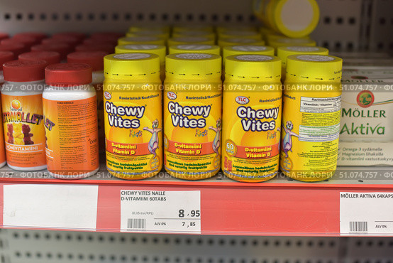 A vitamin store shelf displaying Chewy Vites Kids Vitamin D chewable vitamins. The jars are bright yellow with yellow lids and a colorful label featuring a cartoon character. The front states that the product contains vitamin D, which is good for bone health and immunity, and does not contain artificial flavors. Each package contains 60 chewable tablets with natural fruit flavors. In the foreground is a price tag with a price of 8.95 euros. On the left are other vitamin jars, on the right is a package of Möller Aktiva. The lighting in the store is bright, the photo is taken with the focus on the central packages. (2015 год). Редакционное фото, фотограф Анна Кривицкая / Фотобанк Лори