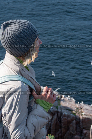 A woman in a gray beanie and light jacket stands on a rocky cliff by the Barents Sea, gazing at the flying seabirds near a nesting colony. Стоковое фото, фотограф Софья Соломонова / Фотобанк Лори