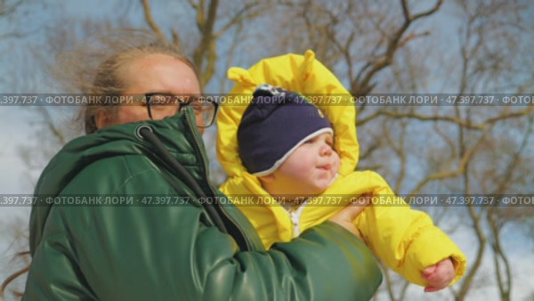 A woman in a green down jacket holds an infant in a bright yellow winter coat against a winter sky. Mother and son on a walk in the park. Close-up, view from below. Стоковое видео, видеограф Воловодов Владимир / Фотобанк Лори