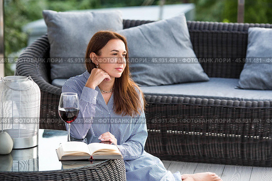 A woman is relaxing on the veranda of a country house and reading a book over a glass of red alcoholic drink or wine. The concept of relaxation and luxury wine. Стоковое фото, фотограф Photojuli / Фотобанк Лори