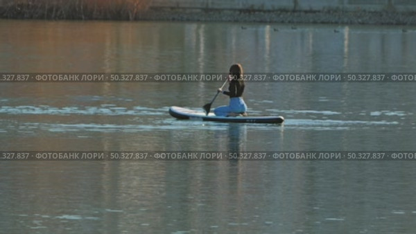 A woman kneeling on a paddleboard and gently paddling into the calm waters of a lake. Dressed in a black top and light blue jeans. Riding a sup board. Russia, January 30, 2025. Редакционное видео, видеограф Воловодов Владимир / Фотобанк Лори