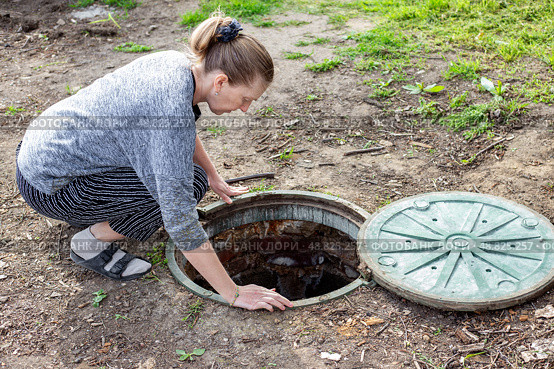 A woman looks into an open septic tank to check the level of sewer discharge. Septic tanks in a country house. Стоковое фото, фотограф Коханова Наталья / Фотобанк Лори