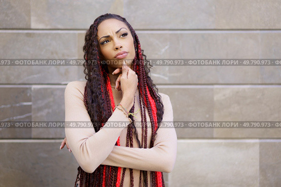 A woman stands thoughtfully against an intriguing textured wall, showcasing her distinctive braided hairstyle adorned with vibrant red accents, embodying confidence, creativity, and personal style. A thoughtful woman sporting braids poses gracefully in a modern urban setting full of life. Стоковое фото, агентство Ingram Publishing / Фотобанк Лори