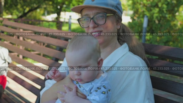 A woman wearing glasses and a baseball cap holds a chubby infant in a patterned jumpsuit, sitting on a wooden bench in a park surrounded by green foliage, on a sunny day. Mother and son on a walk. Стоковое видео, видеограф Воловодов Владимир / Фотобанк Лори
