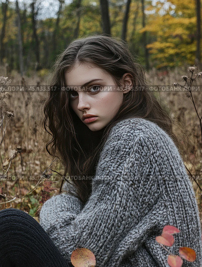 A young girl with long dark hair sitting in an autumn forest. She is wearing a warm gray sweater, which gives the scene a cozy and melancholy atmosphere. The background is blurred, which allows you to focus on the girl and emphasizes her thoughtful look. The nature around shows withering plants and autumn leaves. Стоковое AI-изображение, AI-художник Анна Кривицкая / Фотобанк Лори